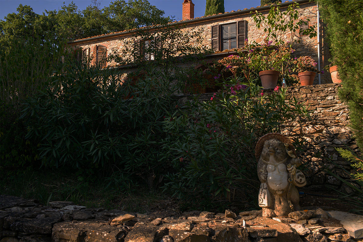Steinernes Landhaus mit Fensterläden, umgeben von dichter Vegetation und einer kleinen Tierstatue im Vordergrund im Abendlicht.